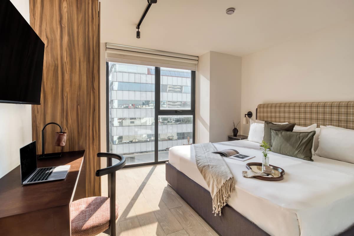 Ein modernes Hotelzimmer mit einem großen Bett, einem Schreibtisch mit einem Laptop, einem Sessel und Blick auf die Stadt durch ein großes Fenster.