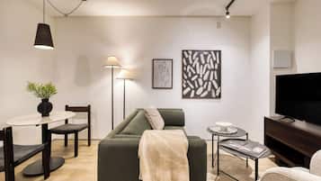 Un salon moderne avec un canapé vert, une table à manger avec des chaises, un lampadaire, un téléviseur et des œuvres d’art accrochées au mur.