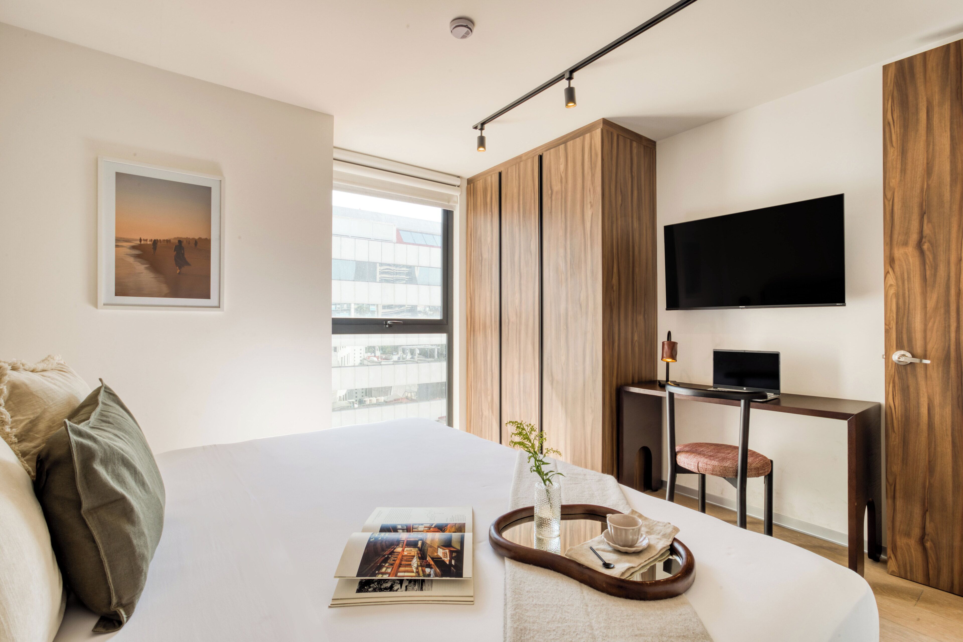 A modern hotel room with a large bed, a desk with a computer, a TV, and a window with a city view.