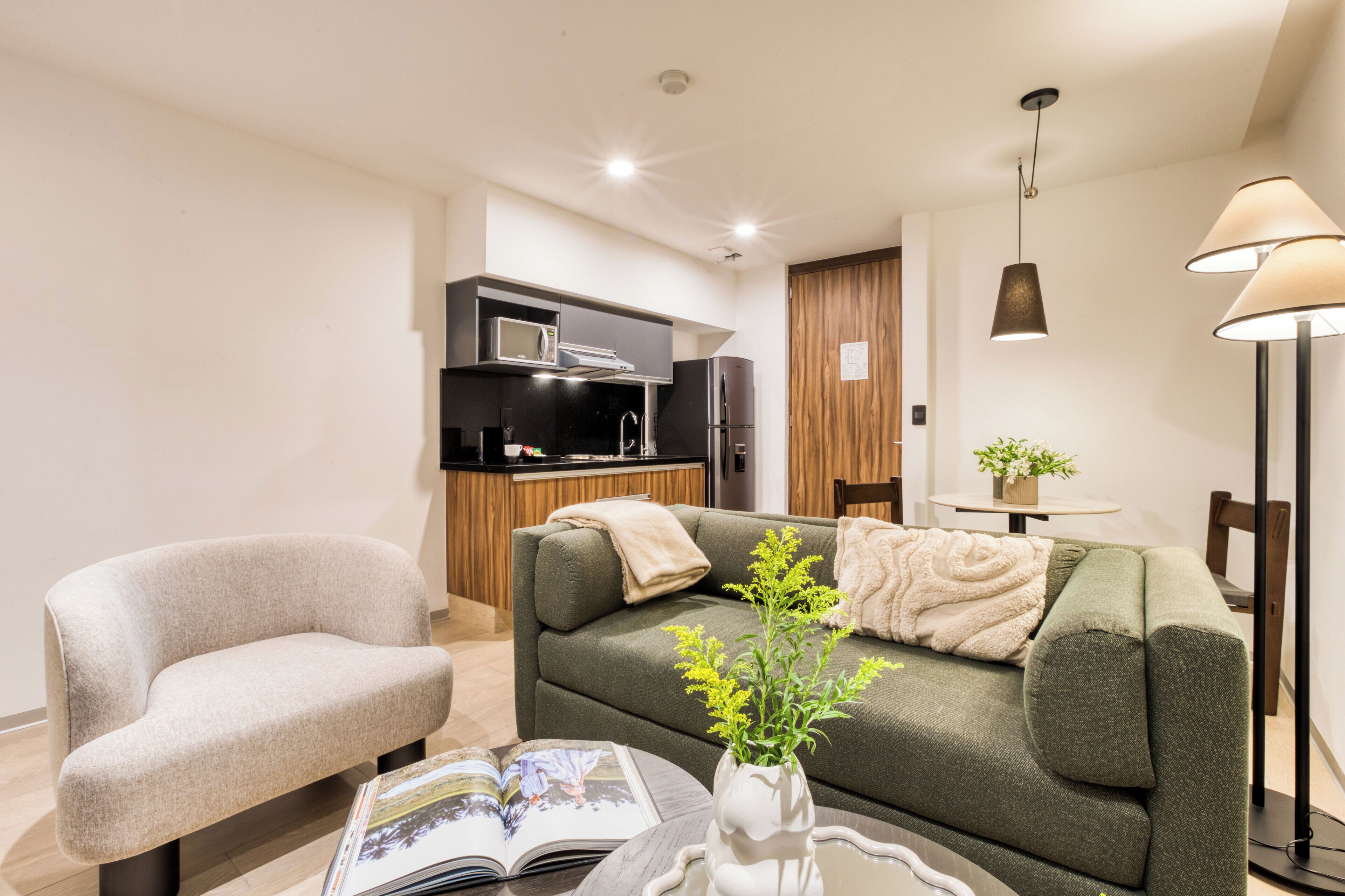 A modern living room with a sofa, a chair, a coffee table with a book, a vase with flowers, and a kitchen in the background.