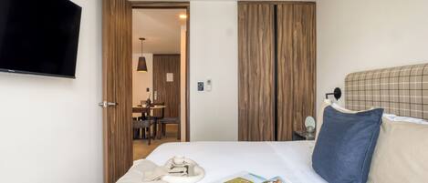 A hotel room with a bed, a flat-screen TV mounted on the wall, a dining area with a table and chairs, and wooden paneling on the walls.