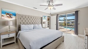 A bedroom with a large bed, a ceiling fan, a nightstand, a lamp, and a view of a swimming pool through a sliding glass door. - Summer Sale! 6 min to Downtown-Waterfront, Heated Pool, Beach Essentials (Cape Coral)