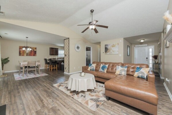 A spacious living room with a brown leather sofa, a dining table, and a ceiling fan.
