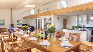 A spacious dining area with a long wooden table set for a meal, flanked by wooden chairs. A kitchen is visible in the background with modern appliances. - Moult Hill Barn (Salcombe)