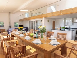Un comedor amplio con una larga mesa de madera dispuesta para una comida, flanqueada por sillas de madera. Se ve una cocina al fondo con electrodomésticos modernos.