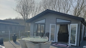 A modern outdoor deck with a glass door leading to a covered patio area, surrounded by a wooden deck and a fenced garden. - 2-bed Lodge, Sleeps 5 w/ Hot Tub & Lake View (Beverley)
