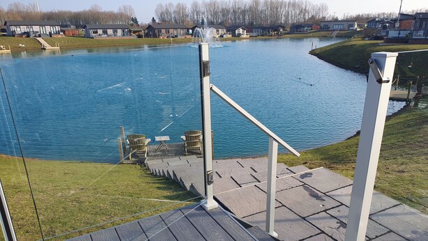 A wooden deck with a railing overlooking a calm lake, with houses in the background. - 2-bed Lodge, Sleeps 5 w/ Hot Tub & Lake View (Beverley)