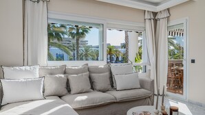 A modern living room with a beige sectional sofa, a round coffee table, and a view of palm trees and buildings through large windows. - Front Line Playa Rocio P. Banus (Marbella)