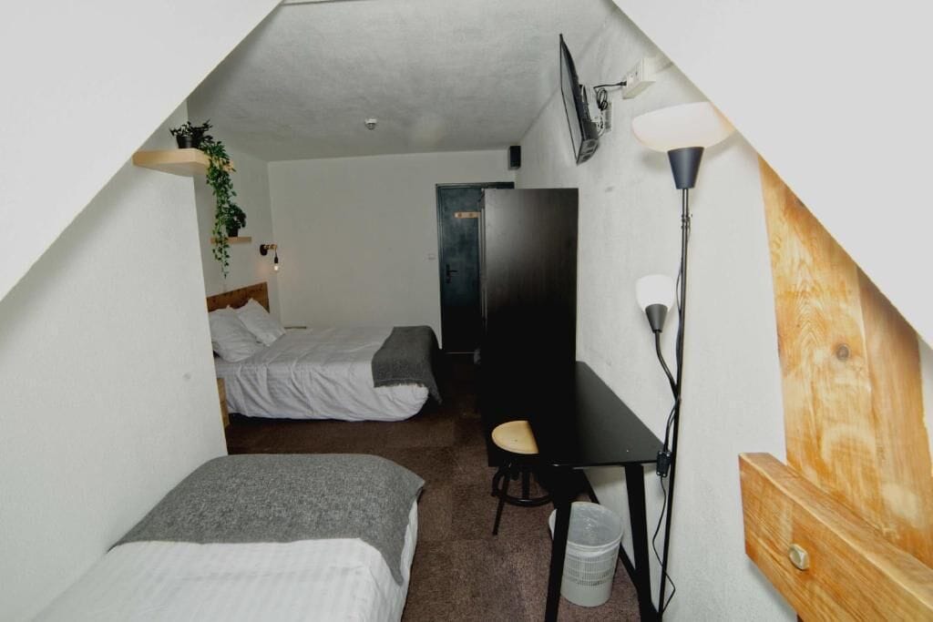 A small, triangular room with two single beds, a desk, a chair, a TV, and a lamp.