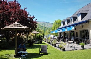 An outdoor cafe area with tables and chairs, a thatched umbrella, and a building with a blue awning.