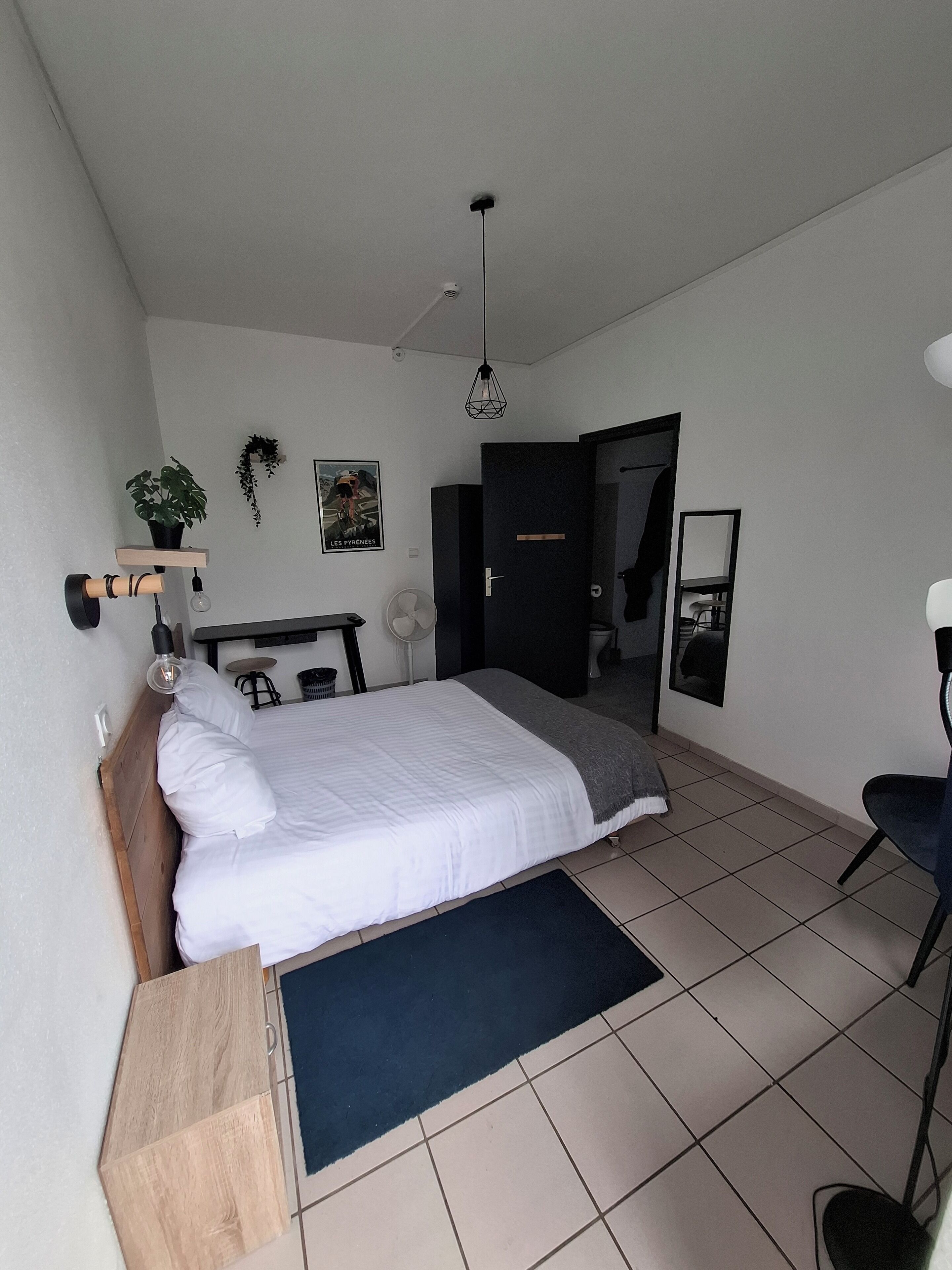 A modern bedroom with a bed, a desk, a chair, a fan, and a potted plant.