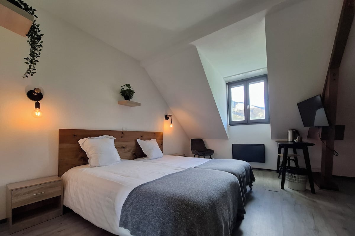 A bedroom with a sloped ceiling, a bed with white linens, a chair, a desk with a computer, and a window with a view.