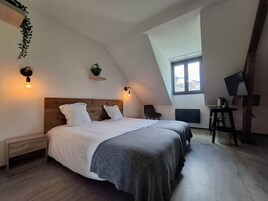 A bedroom with a sloped ceiling, a bed with white linens, a chair, a desk with a computer, and a window with a view.