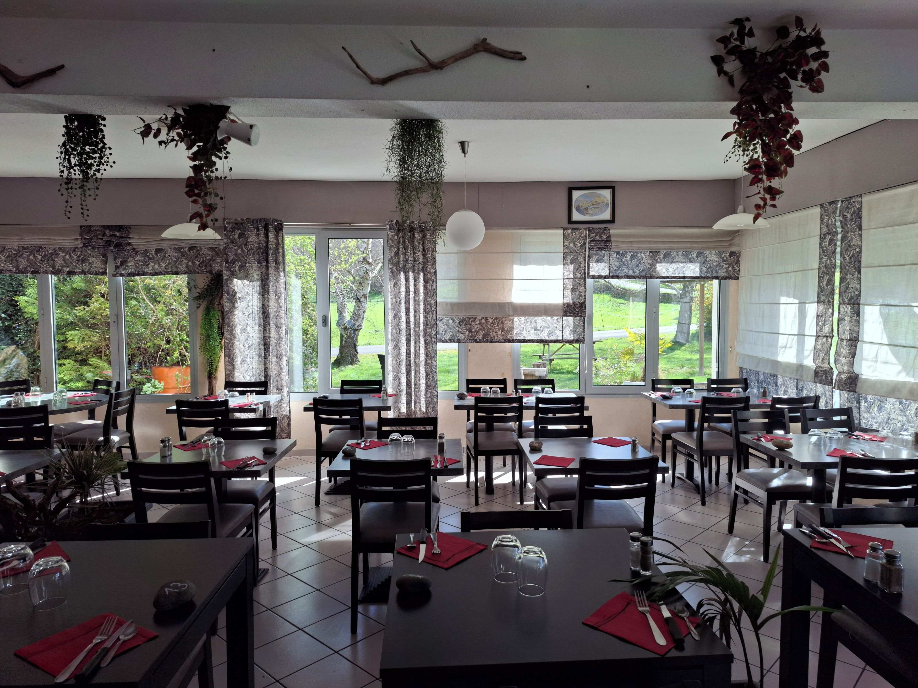 A restaurant with multiple tables and chairs, large windows with a view of greenery, and hanging plants.