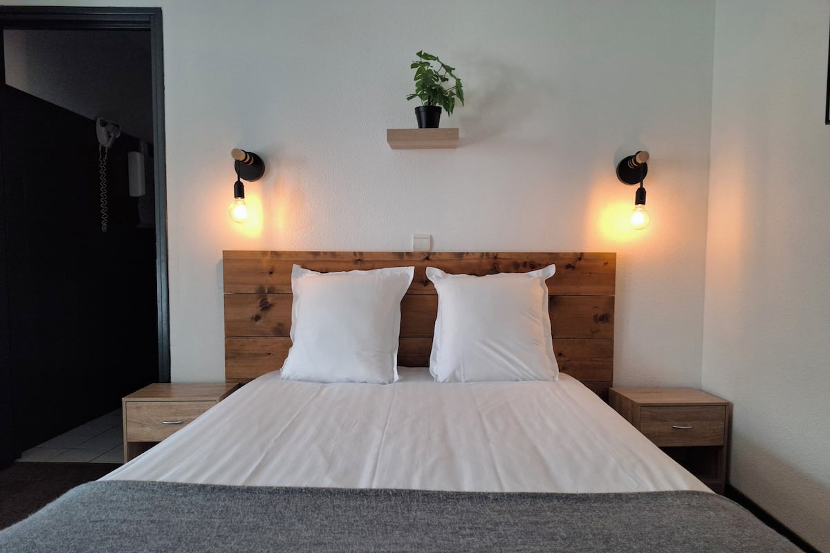 A neatly made bed with white linens and two pillows, flanked by wooden nightstands with black wall sconces, a small wooden shelf with a potted plant, and a dark doorway to the left.