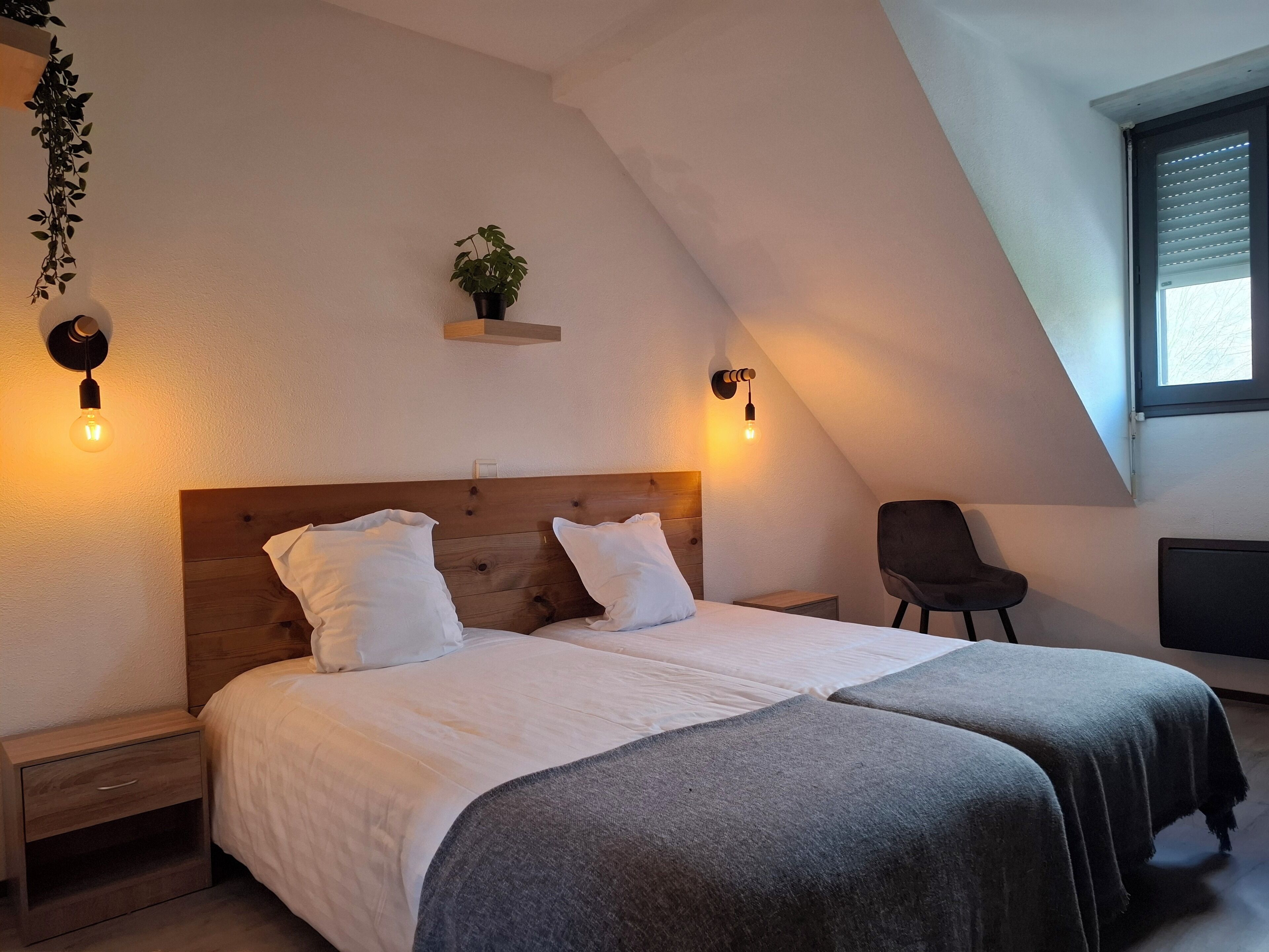 Un dormitorio con cabecera de madera, dos camas, mesitas de noche, una silla, una planta y una ventana con persianas.