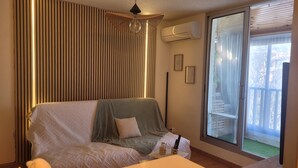 A bedroom with a bed, a bedside table, a hanging light fixture, a window with a view, and an air conditioning unit.