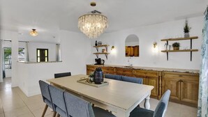 A spacious dining area with a large table and chairs, a chandelier, wooden cabinets, and built-in shelves with decorative items. - Welcome To The Leaf House (Delray Beach)