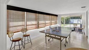 A ping pong table with chairs, large windows with blinds, and a view of the outside. - Welcome To The Leaf House (Delray Beach)