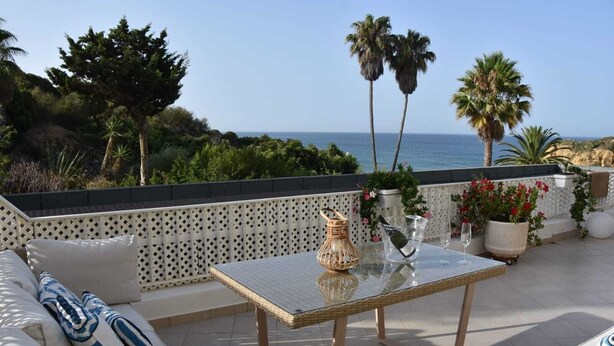 A terrace with a glass table, white cushioned seating, and potted plants. - NEW apt. 1 min. walk from beach, sea view, large terrace, two pools (Albufeira)