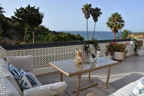 Eine Terrasse mit Glastisch, weiß gepolsterten Sitzmöglichkeiten und Topfpflanzen.