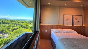 Quarto com cama, janela com vista para vegetação e obras de arte emolduradas na parede.