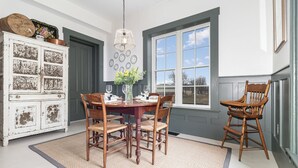 A dining area with a wooden table set for four, surrounded by chairs, a white cabinet with decorative items, and a large window with a view. - Historic Home & Farm in Bourbon Country  (Warsaw)