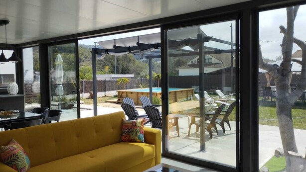 A modern living room with a yellow sofa, a glass coffee table, and large windows offering a view of a backyard with a pool and outdoor furniture.