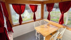 Una veranda con un tavolo di legno e sedie bianche, ampie finestre con tende rosse e vista su un giardino.