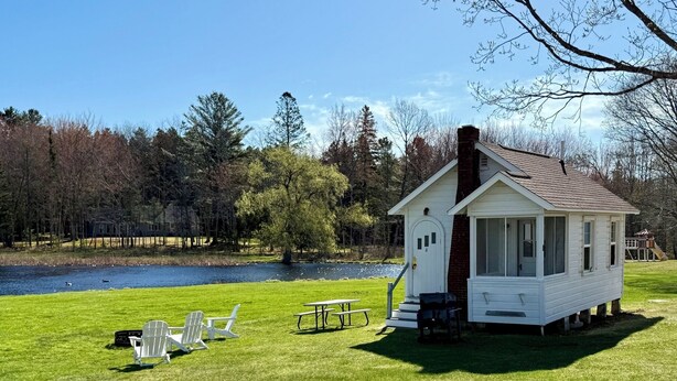 A small white cabin with a chimney, a picnic table, and two chairs on a grassy area by a lake. - Bar Harbor Cottage Close to Acadia & Downtown Bar Harbor - Kitchen, AC, Wi-Fi, Fireplace (Sleeps 6). (Bar Harbor)