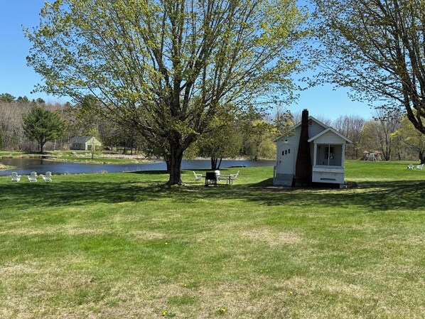 A small cabin with a chimney, a picnic table, and a bench near a pond.