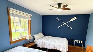 A bedroom with two beds, a ceiling fan, a window with blinds, and a wall-mounted cross symbol.