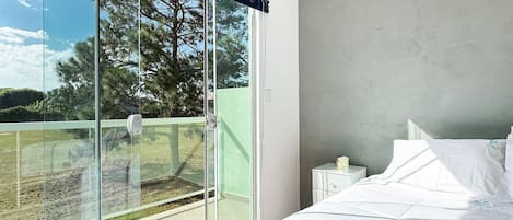 A bedroom with a large sliding glass door leading to a balcony, a bed with white bedding, and a nightstand with a lamp.