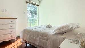 A bedroom with a bed, a dresser, and a window with a view of trees. - Casa de Andréa com piscina - Campeche (Florianopolis)