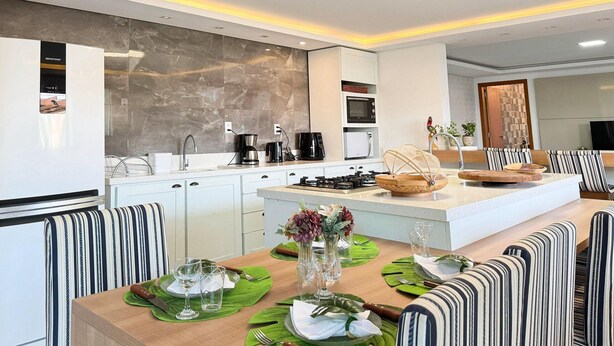 A modern kitchen with a central island, white cabinets, and a dining area with striped chairs and a wooden table set for a meal. - Casa de Andréa com piscina - Campeche (Florianopolis)