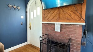 A room with a blue wall, a wooden door, a TV mounted on a wooden wall, and a black metal rack. - Studio cottage in Bar Harbor - Cozy 1930's Fireplace, Sleeps 3. (Bar Harbor)