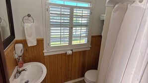 A bathroom with a wooden floor, white fixtures, a window with blinds, and a shower curtain. - Studio cottage in Bar Harbor - Cozy 1930's Fireplace, Sleeps 3. (Bar Harbor)