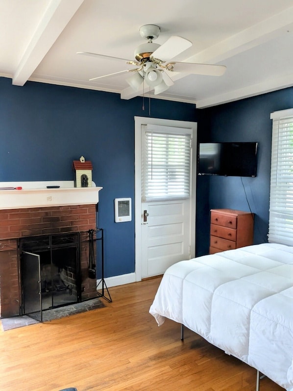 A bedroom with a white bed, a ceiling fan, a fireplace, a TV, and a door with blinds.