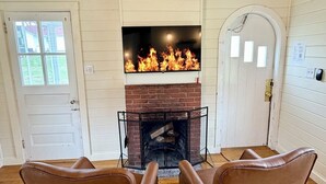 A room with a fireplace, two leather chairs, wooden flooring, and a white door. - Cozy studio cottage in Bar Harbor - Kitchen, AC, Wi-Fi & Fireplace (Bar Harbor)