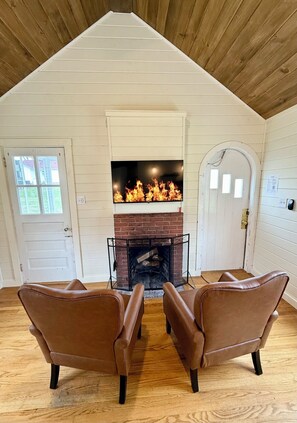 A room with a fireplace, two leather chairs, wooden flooring, and a white door. - Cozy studio cottage in Bar Harbor - Kitchen, AC, Wi-Fi & Fireplace (Bar Harbor)
