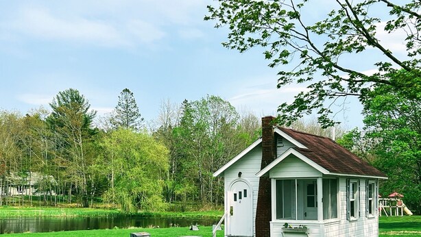A small white cabin with a chimney, a porch, and a picnic table set on a grassy area near a body of water. - Cozy studio cottage in Bar Harbor - Kitchen, AC, Wi-Fi & Fireplace (Bar Harbor)