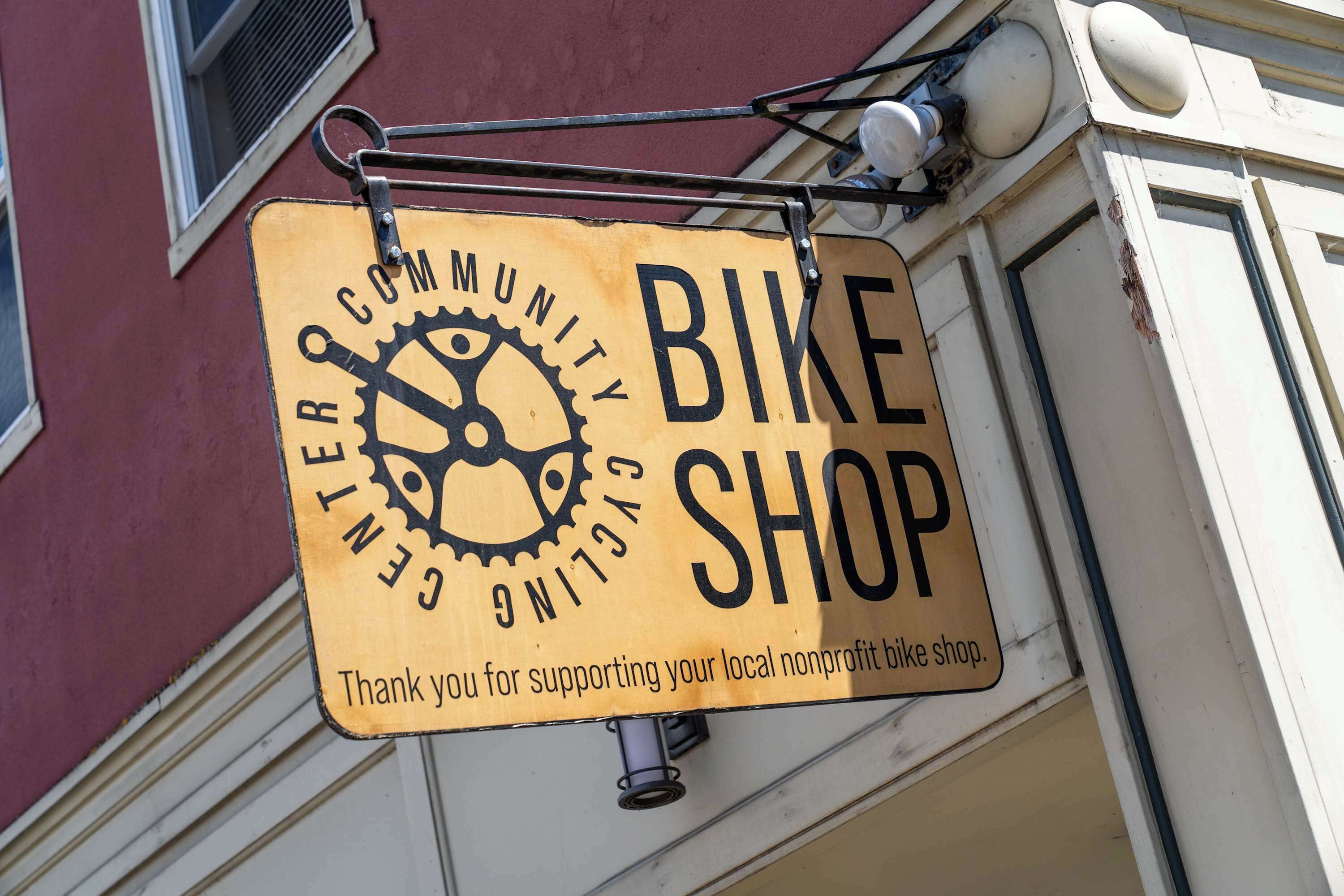 A yellow sign with black text hanging from a building, reading "Community Gear Cycling Shop."