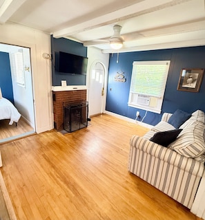 A room with a wooden floor, a striped sofa, a television, a fireplace, and a window with blinds.