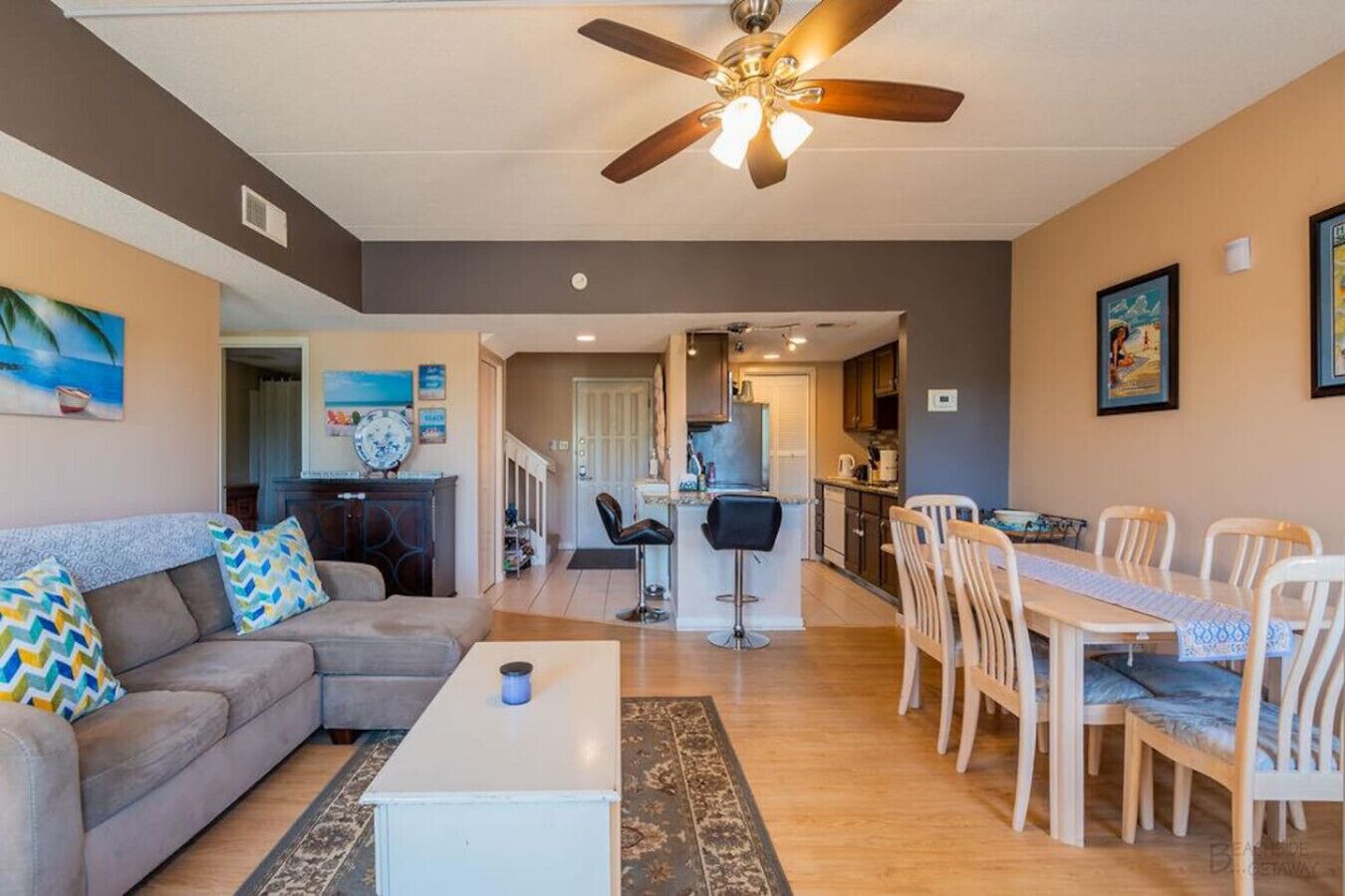 A living room with a sofa, coffee table, and dining area with chairs and a table. There is a ceiling fan and a kitchen in the background.