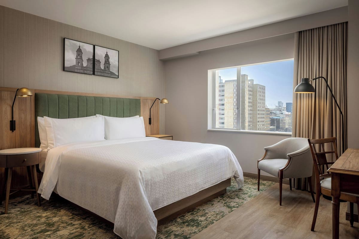 Une chambre d’hôtel avec un grand lit, deux tables de chevet, une chaise, une petite table et une fenêtre donnant sur la ville.