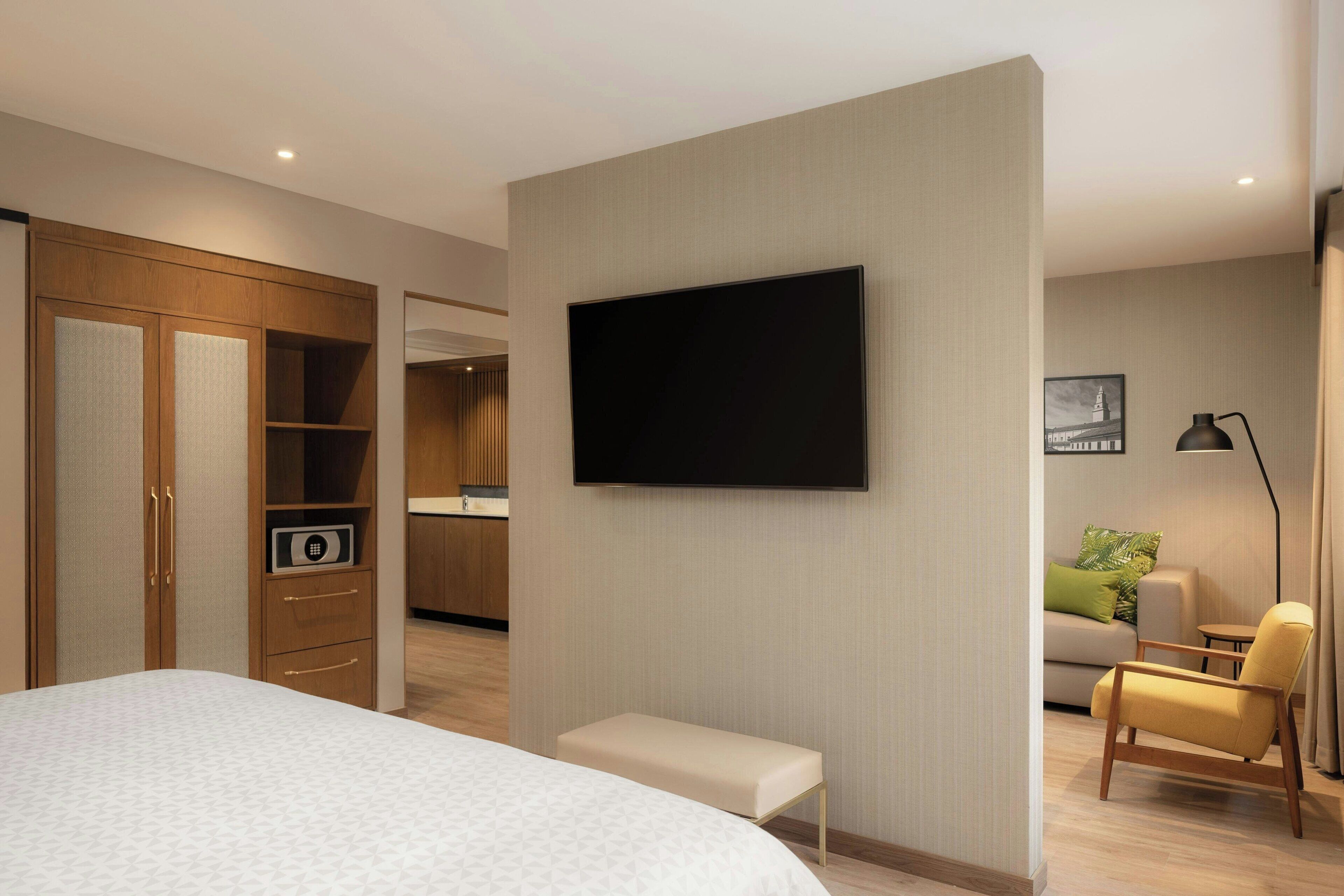 A modern hotel room with a large bed, a TV mounted on the wall, a small bench, a sofa, and a desk with a lamp.