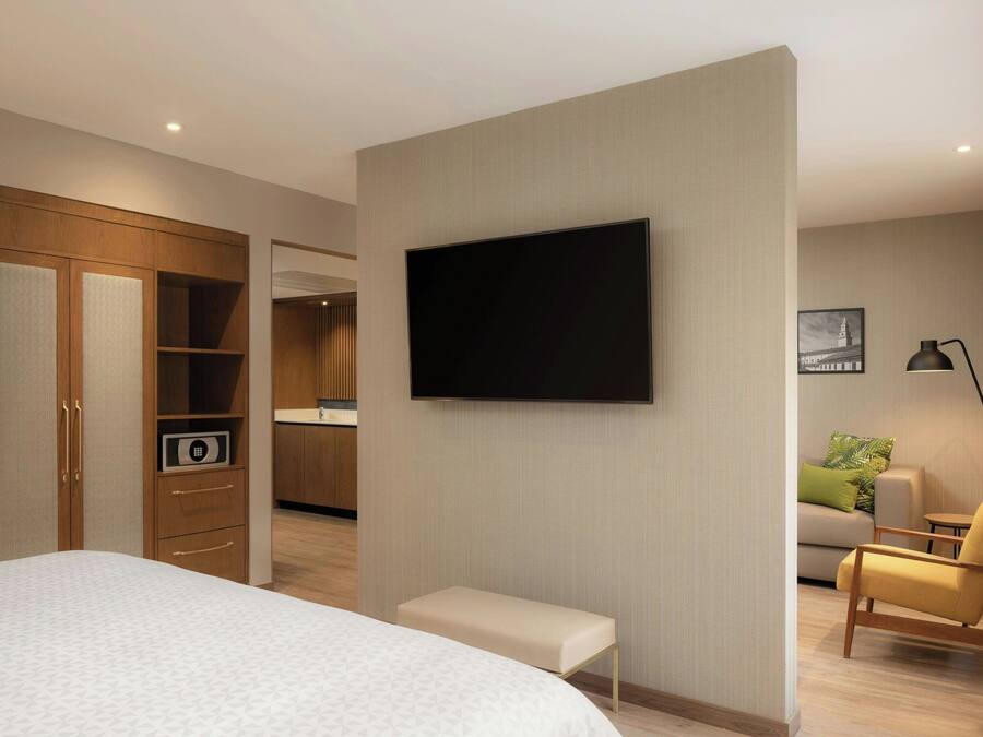 Habitación de hotel moderna con una cama grande, un televisor montado en la pared, un pequeño banco, un sofá y un escritorio con una lámpara.