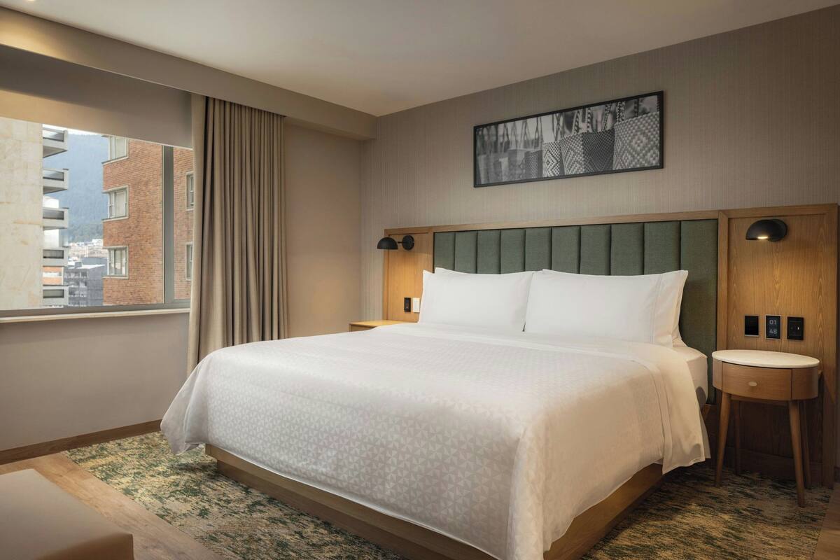 Une chambre d’hôtel avec un grand lit, une table de chevet, une petite table, une fenêtre avec des rideaux et une vue sur la ville.