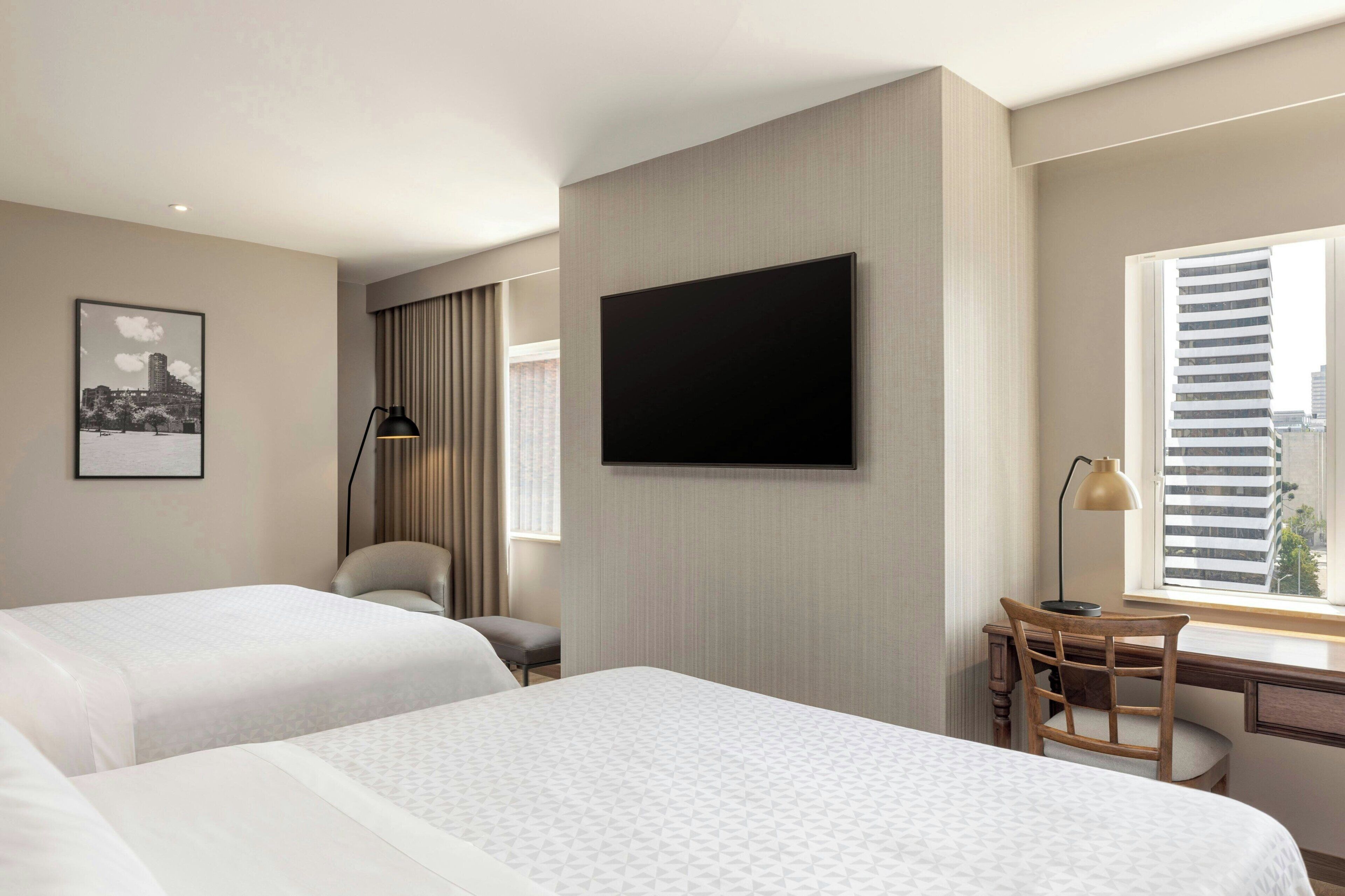 A hotel room with two beds, a desk, a chair, a TV mounted on the wall, and a window with blinds.
