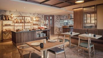 A modern restaurant with a central counter displaying various breads and pastries, surrounded by wooden tables and chairs.